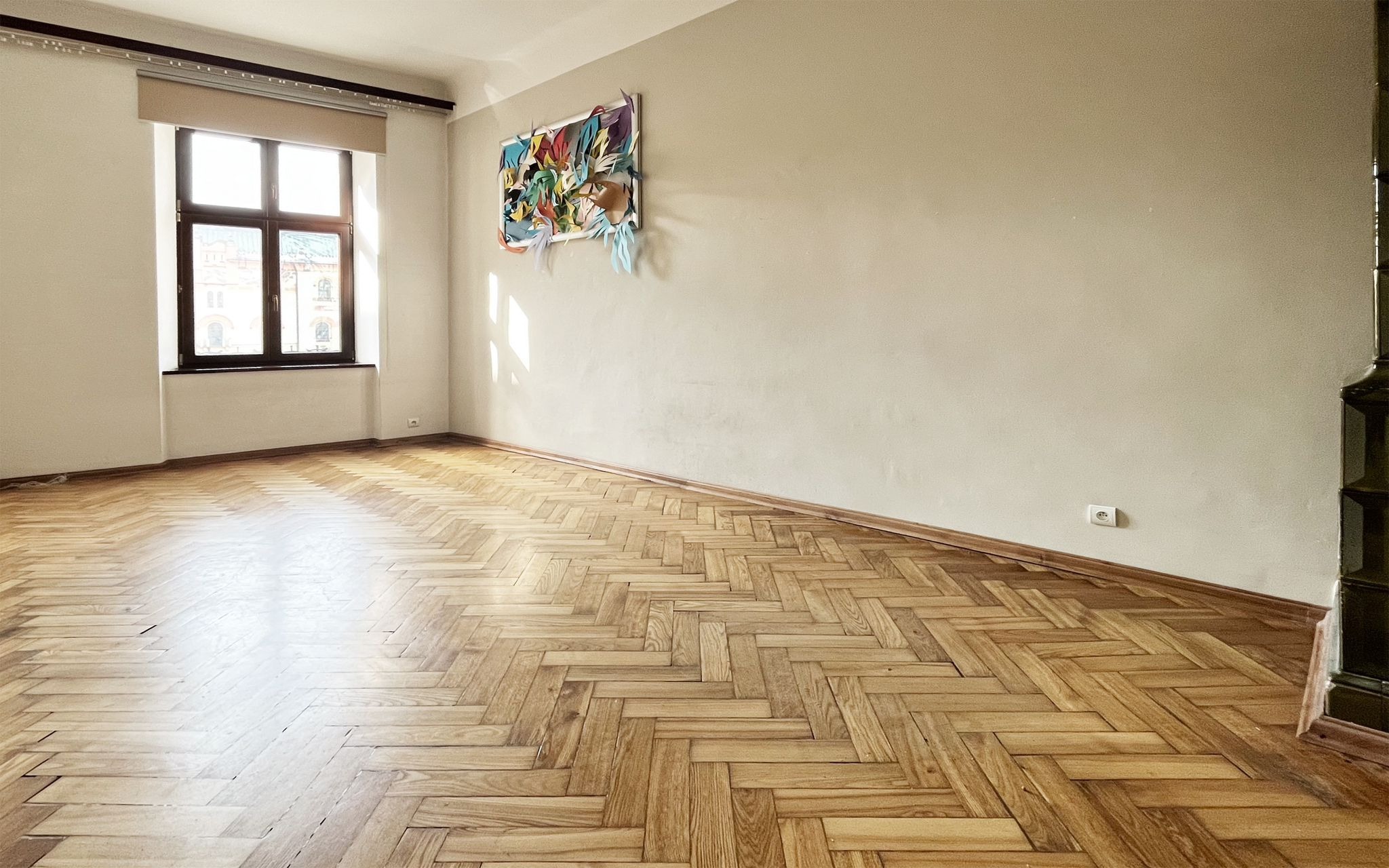 Puste, jasne pomieszczenie z parkietem ułożonym w jodełkę, oknem z widokiem na budynek i abstrakcyjną dekoracją ścienną z papieru.