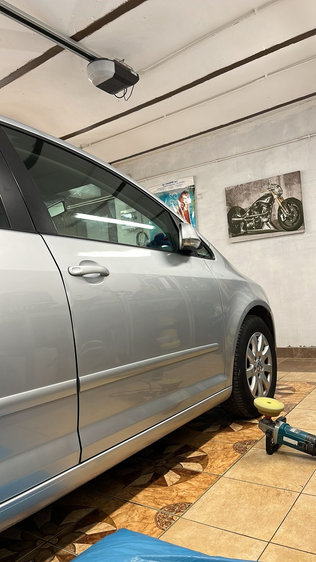 Srebrny samochód w garażu podczas polerowania karoserii, widoczna polerka i pady polerskie, w tle plakat z motocyklem.