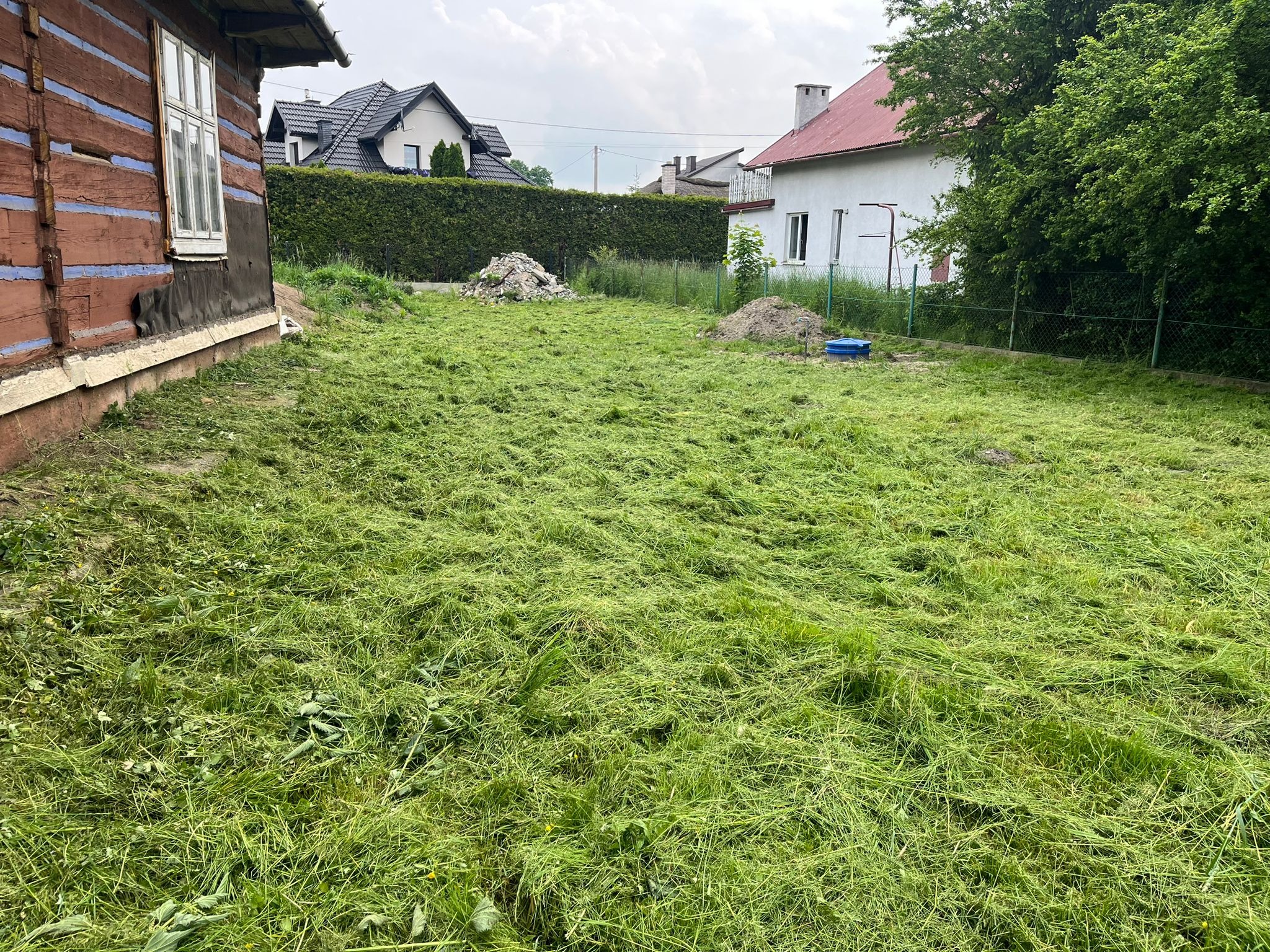 Świeżo skoszona trawa na działce z widocznymi dwoma starymi domami i nowym domem w tle, pozostałościami gruzu i ziemi.