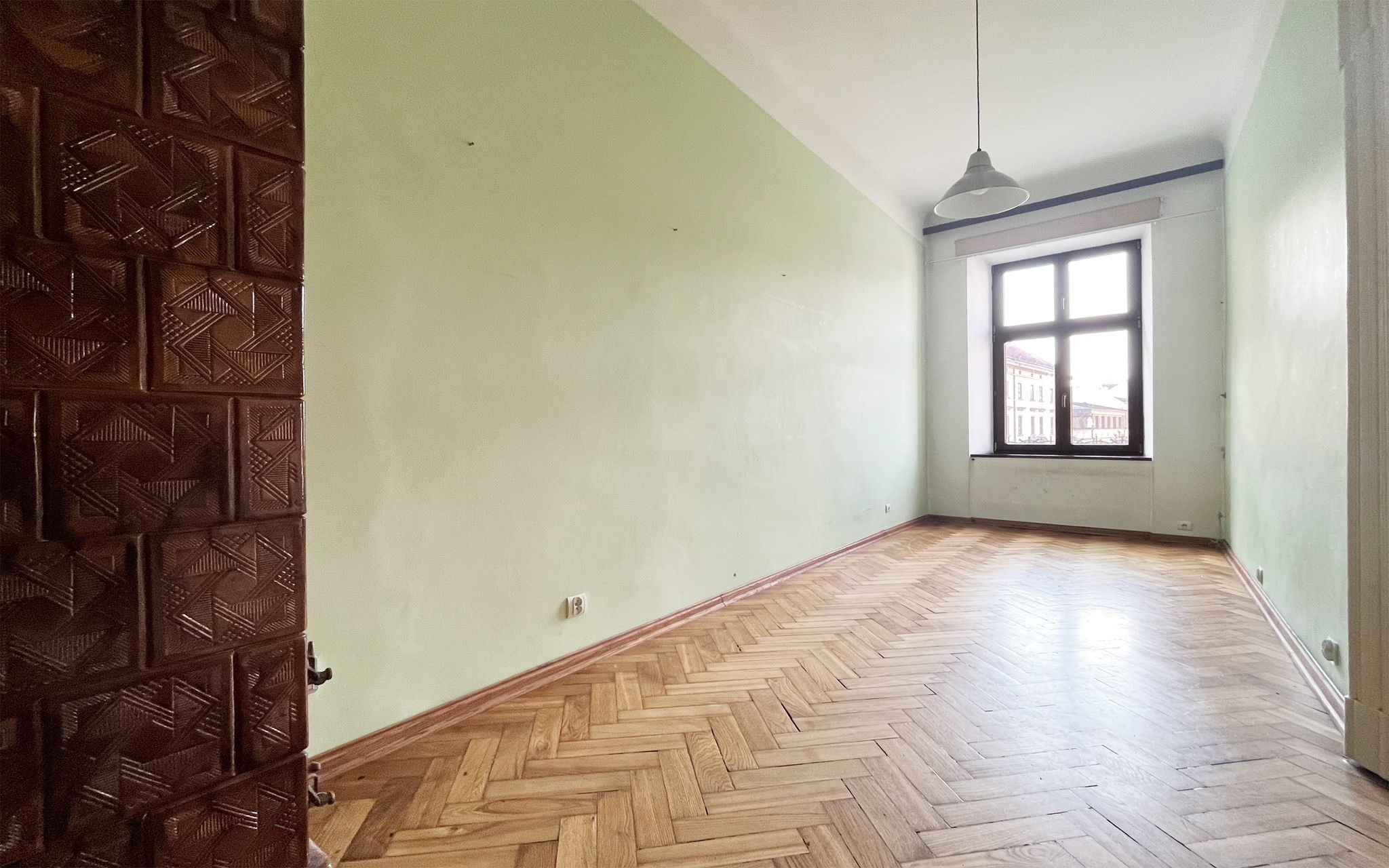 Puste wnętrze pokoju z parkietem ułożonym w jodełkę, piecem kaflowym w rogu i oświetleniem w postaci wiszącej lampy. Widoczne ślady po niedawno usuniętych elementach na ścianach.