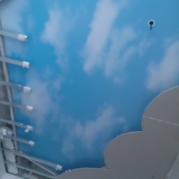 VIP Bud - Nisko kątowe ujęcie sufitu podwieszanego z nadrukiem błękitnego nieba i chmur, widoczne metalowe profile konstrukcyjne i fragment dekoracyjnej płyty kartonowo-gipsowej w kształcie chmury.