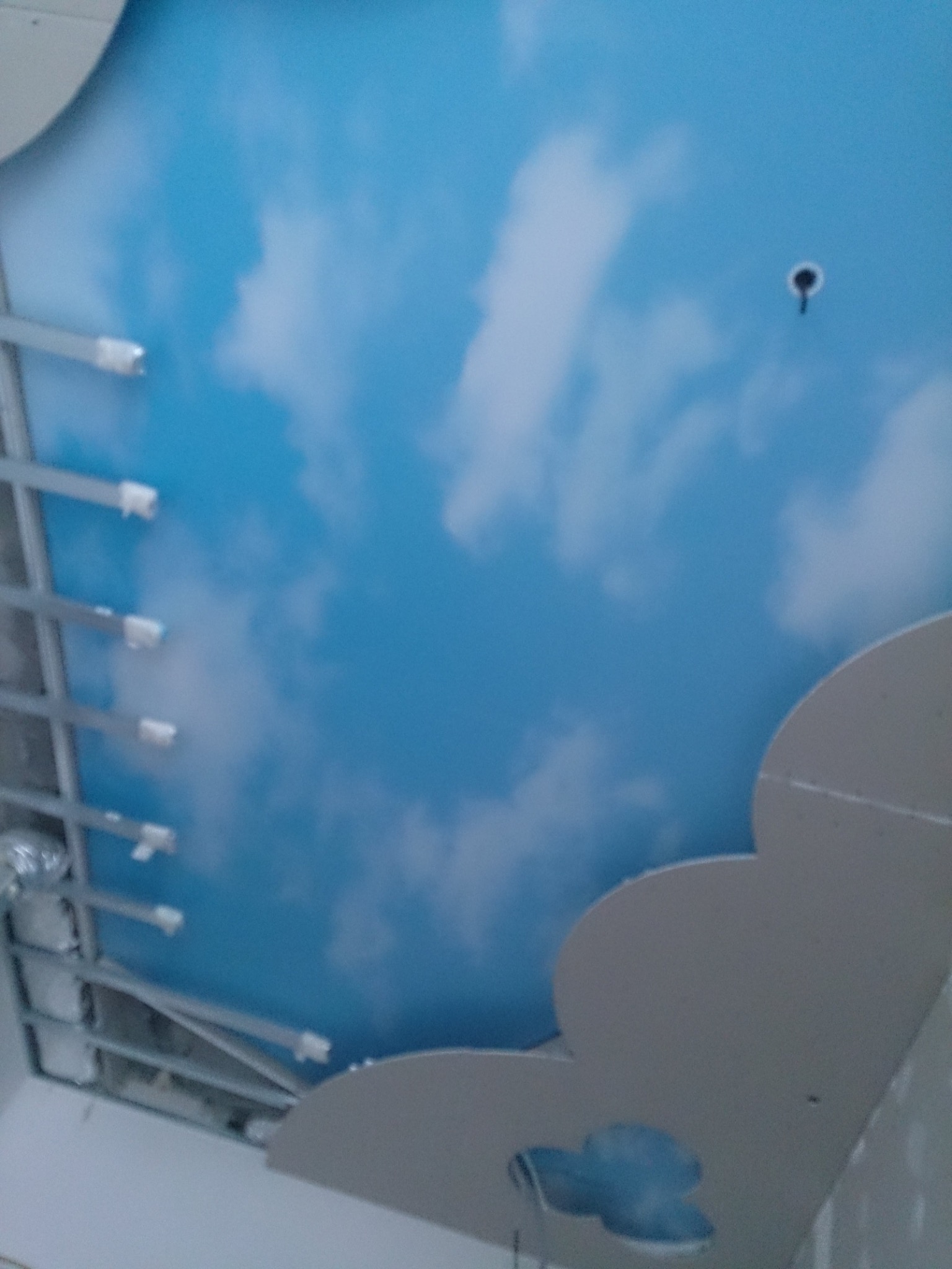 Nisko kątowe ujęcie sufitu podwieszanego z nadrukiem błękitnego nieba i chmur, widoczne metalowe profile konstrukcyjne i fragment dekoracyjnej płyty kartonowo-gipsowej w kształcie chmury.