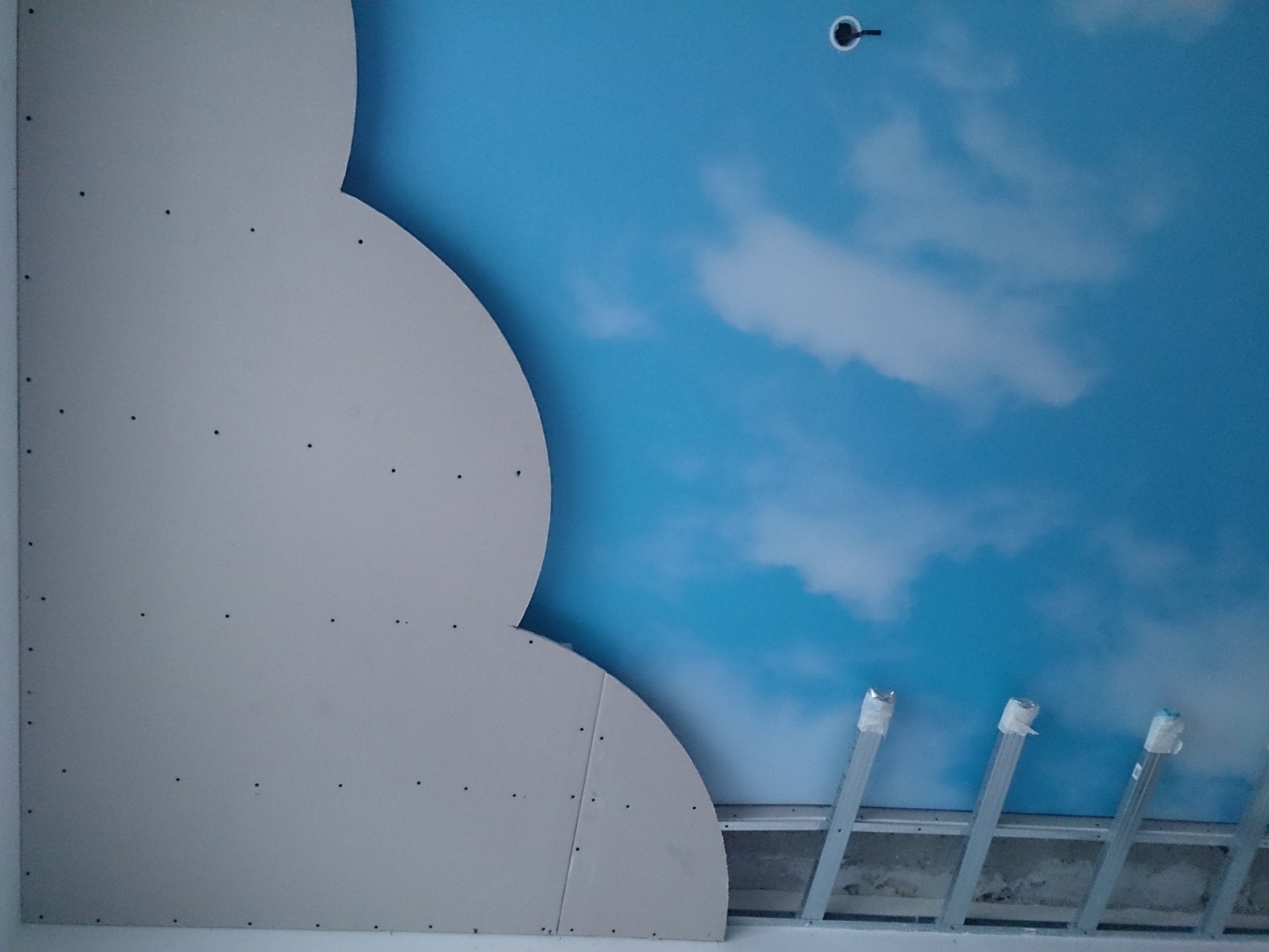 Montaż sufitu napinanego z motywem nieba i chmur, widoczne profile i płyta karton gips w trakcie instalacji.