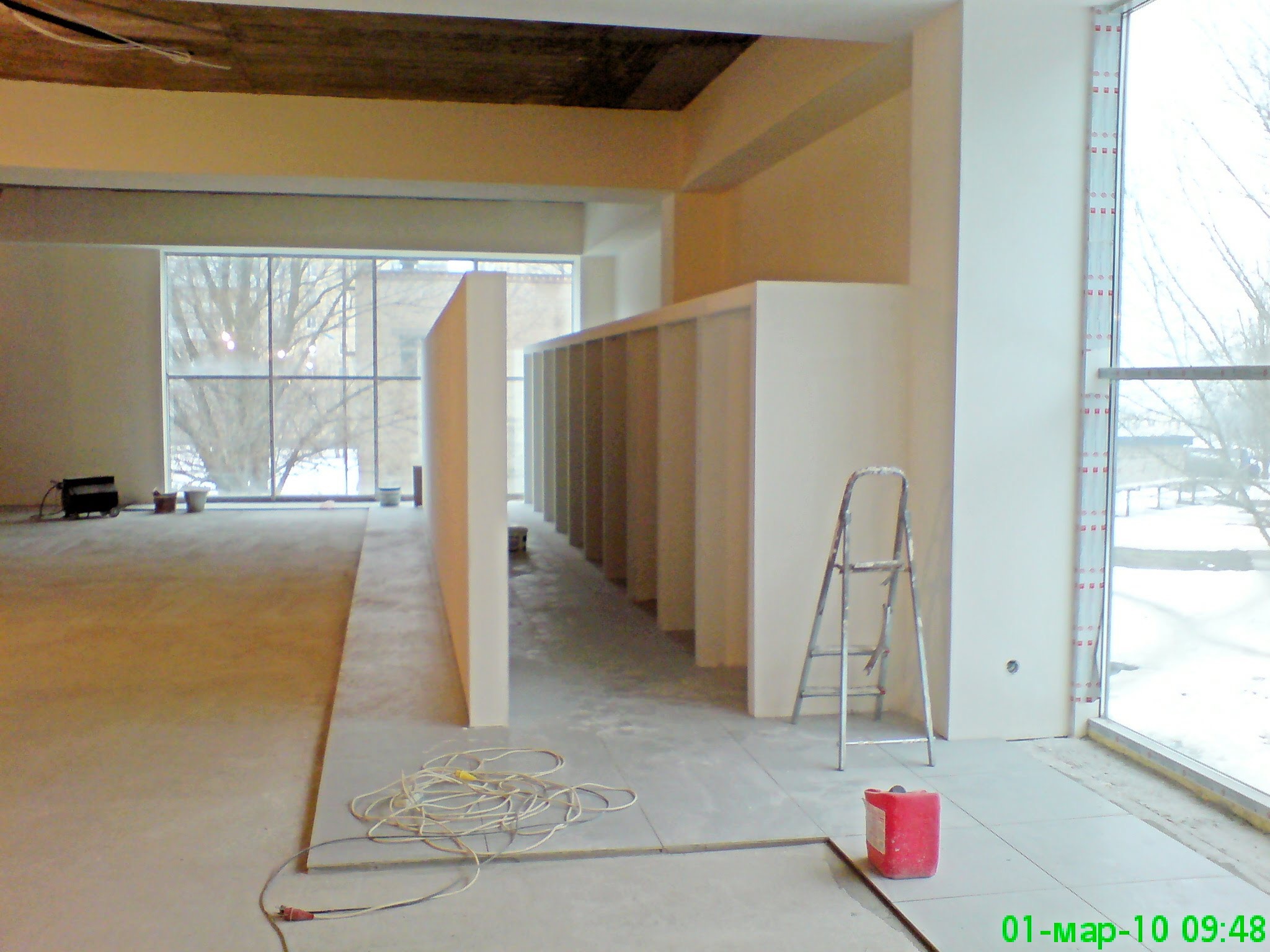 Wnętrze w trakcie prac wykończeniowych: widoczne ściany działowe z płyt kartonowo-gipsowych, częściowo ułożona podłoga, drabina i zwój kabla na posadzce, duże okna z widokiem na zewnątrz.