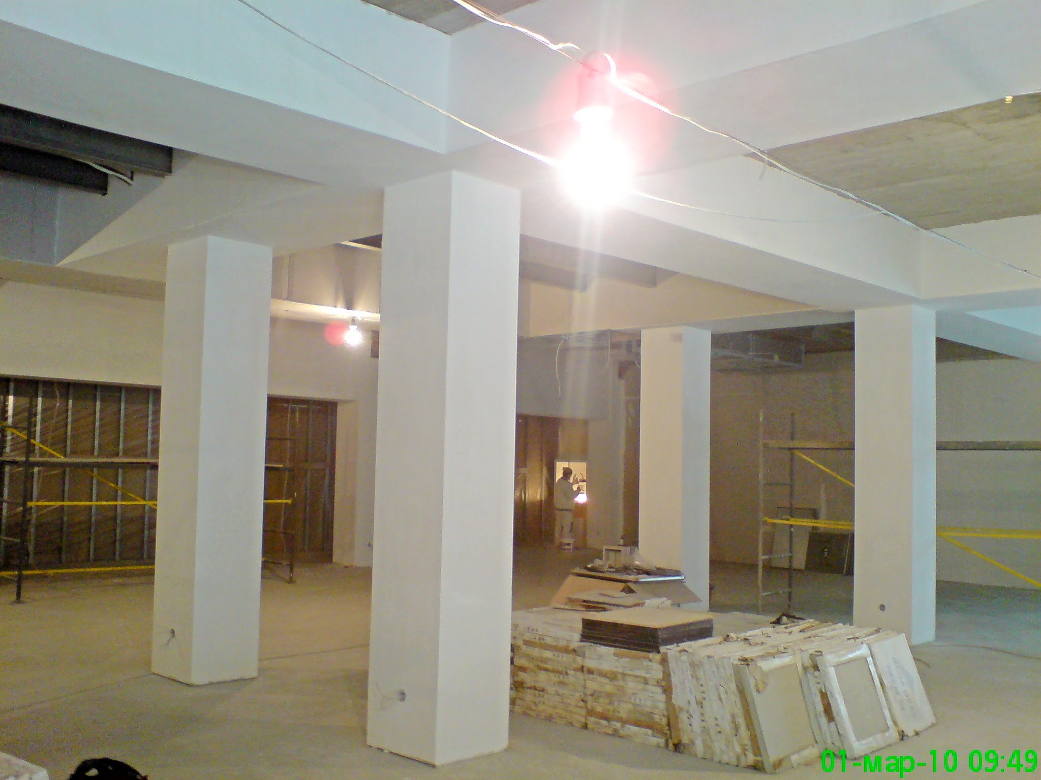 Wnętrze w trakcie prac wykończeniowych: widoczne kolumny konstrukcyjne, surowy betonowy strop, metalowe profile ścian działowych oraz materiały budowlane przygotowane do montażu.