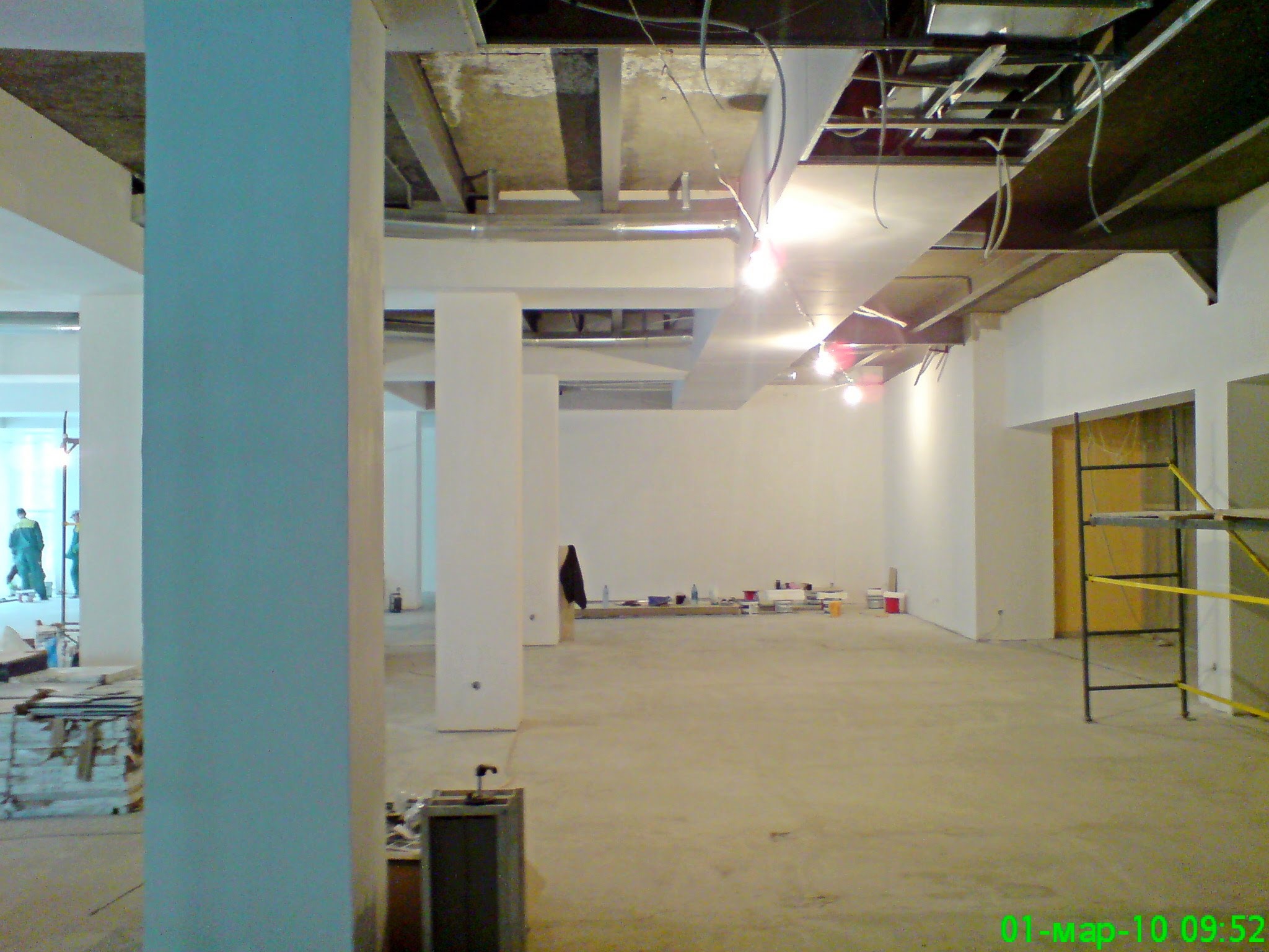Wnętrze w trakcie remontu z widocznymi surowymi stropami, instalacjami pod sufitem, białymi ścianami, kolumnami oraz rozstawionym rusztowaniem, z materiałami budowlanymi rozłożonymi na podłodze.