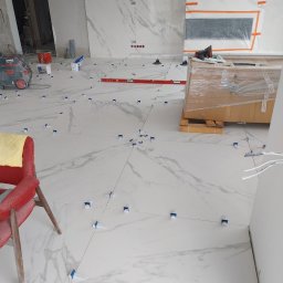 Damian Kański - Białe płytki imitujące marmur układane na podłodze z użyciem poziomicy i klipsów poziomujących, w tle widoczny odkurzacz przemysłowy i zabezpieczony kartonami otwór na kominek.
