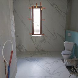 Damian Kański - Łazienka w trakcie układania płytek imitujących marmur na ścianach i podłodze, z oknem zabezpieczonym taśmą malarską, widoczna toaleta i fragmenty instalacji hydraulicznej w surowym stanie.