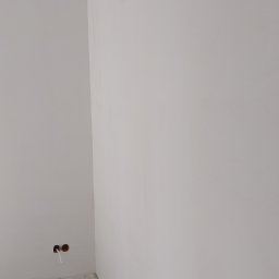 Damian Kański - Wnętrze pokoju w trakcie prac wykończeniowych: ściany pomalowane na biało, sufit z widocznymi miejscami po szpachlowaniu, betonowa podłoga z drobnymi zabrudzeniami, widoczne otwory na gniazdka...