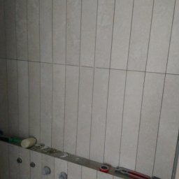 Damian Kański - Ściana łazienki w trakcie wykańczania z ułożonymi pionowo jasnymi płytkami, widoczne narzędzia i elementy instalacji hydraulicznej.