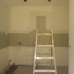 Damian Kański - Łazienka w trakcie wykańczania z częściowo położonymi heksagonalnymi płytkami na ścianie, widoczna drabina malarska i narzędzia do cięcia płytek.