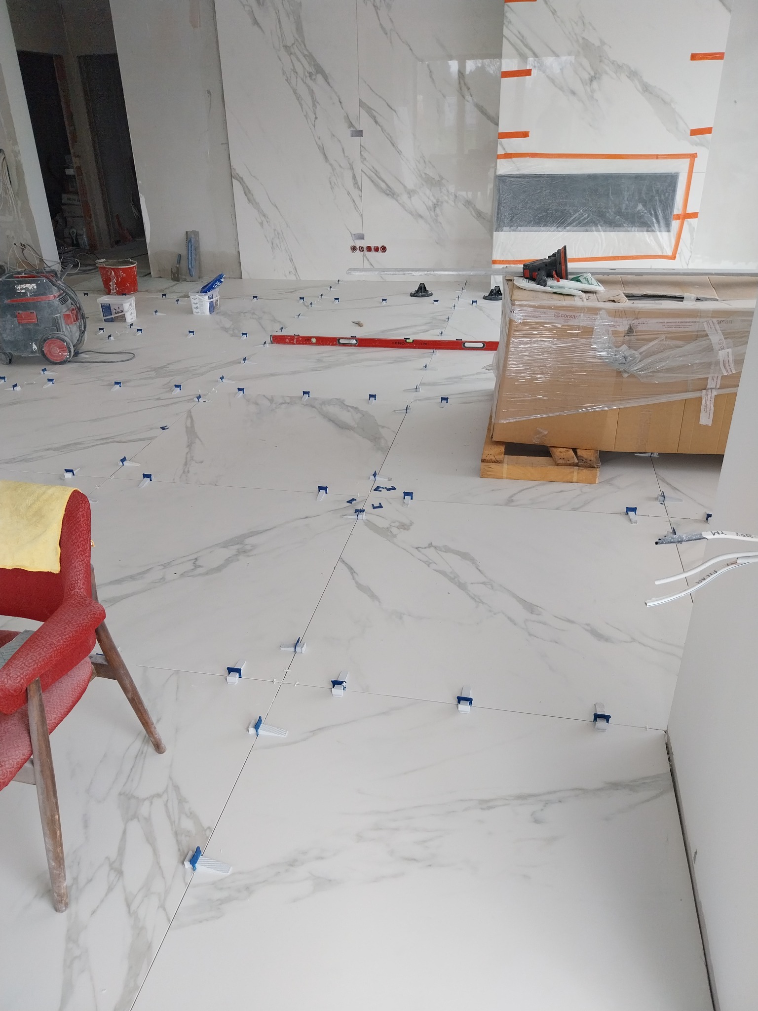 Białe płytki imitujące marmur układane na podłodze z użyciem poziomicy i klipsów poziomujących, w tle widoczny odkurzacz przemysłowy i zabezpieczony kartonami otwór na kominek.