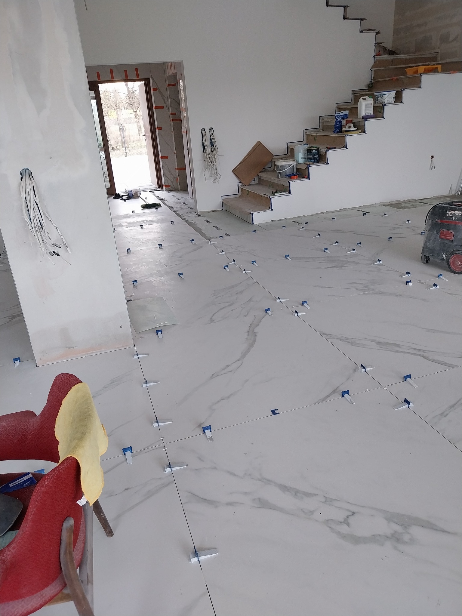 Układanie płytek imitujących marmur na podłodze, z użyciem klinów poziomujących; widoczne schody w stanie surowym, narzędzia oraz odkurzacz przemysłowy.