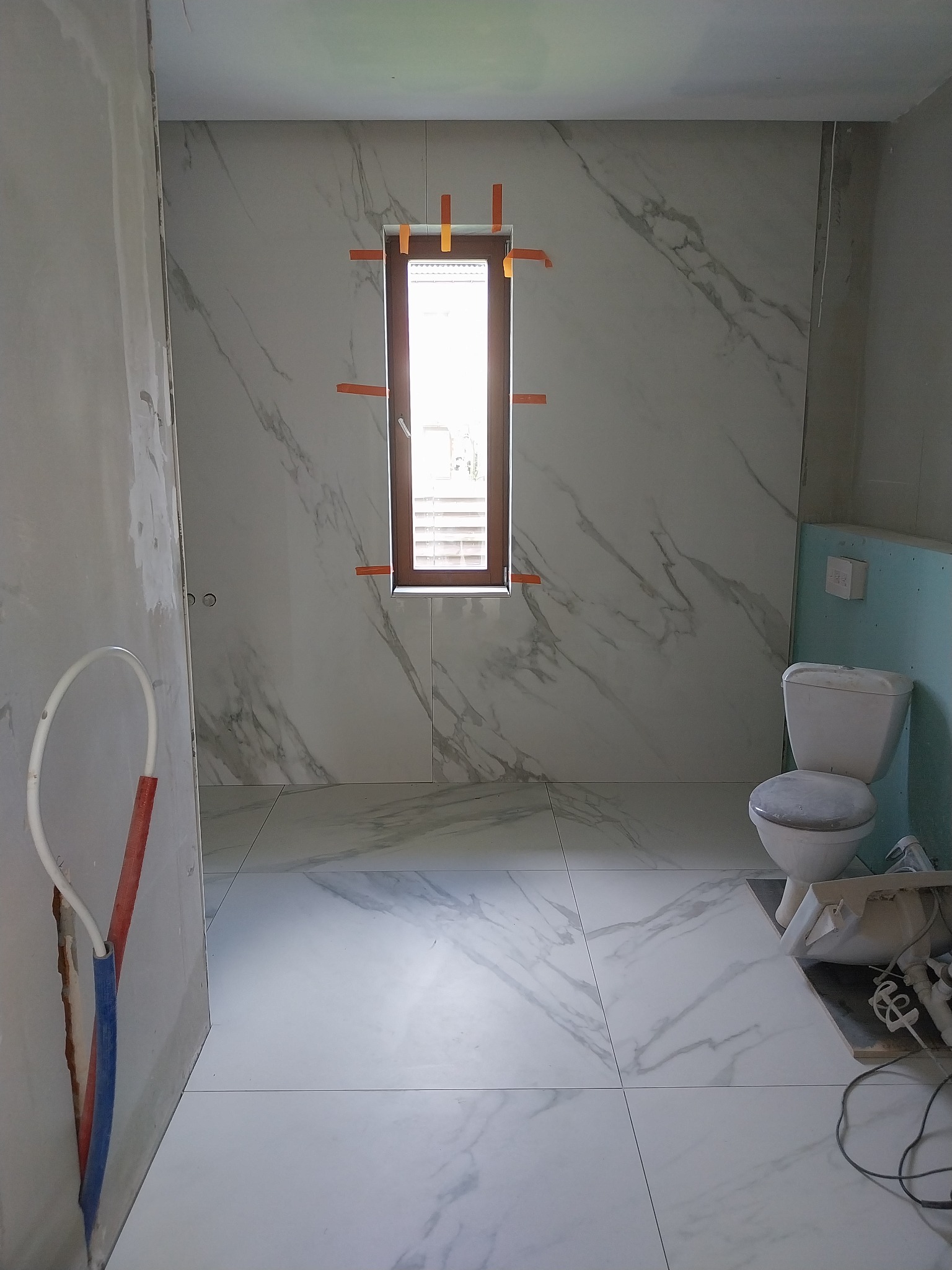 Łazienka w trakcie układania płytek imitujących marmur na ścianach i podłodze, z oknem zabezpieczonym taśmą malarską, widoczna toaleta i fragmenty instalacji hydraulicznej w surowym stanie.
