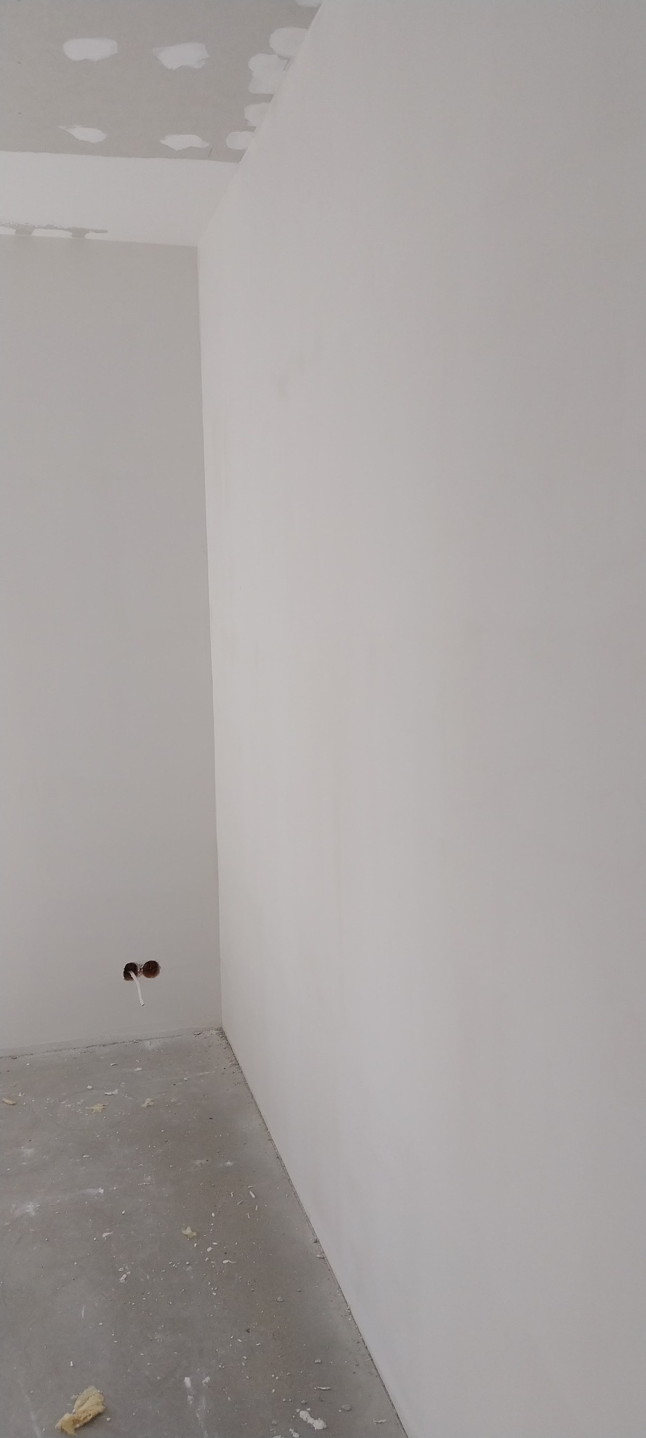 Wnętrze pokoju w trakcie prac wykończeniowych: ściany pomalowane na biało, sufit z widocznymi miejscami po szpachlowaniu, betonowa podłoga z drobnymi zabrudzeniami, widoczne otwory na gniazdka...