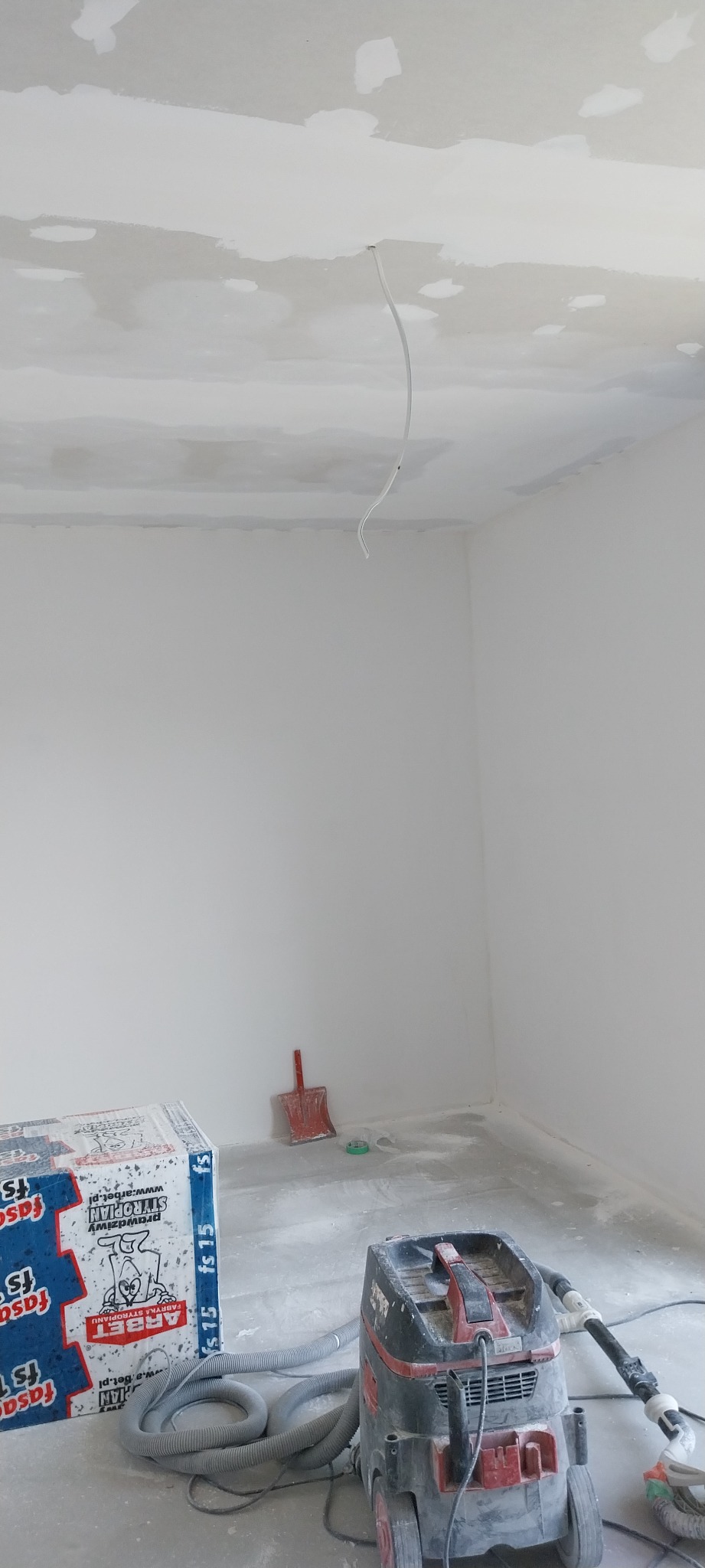 Pokój w trakcie prac wykończeniowych: sufit z widocznymi łatami, wystający kabel, paczka styropianu i odkurzacz przemysłowy, czerwona szpachelka przy ścianie.