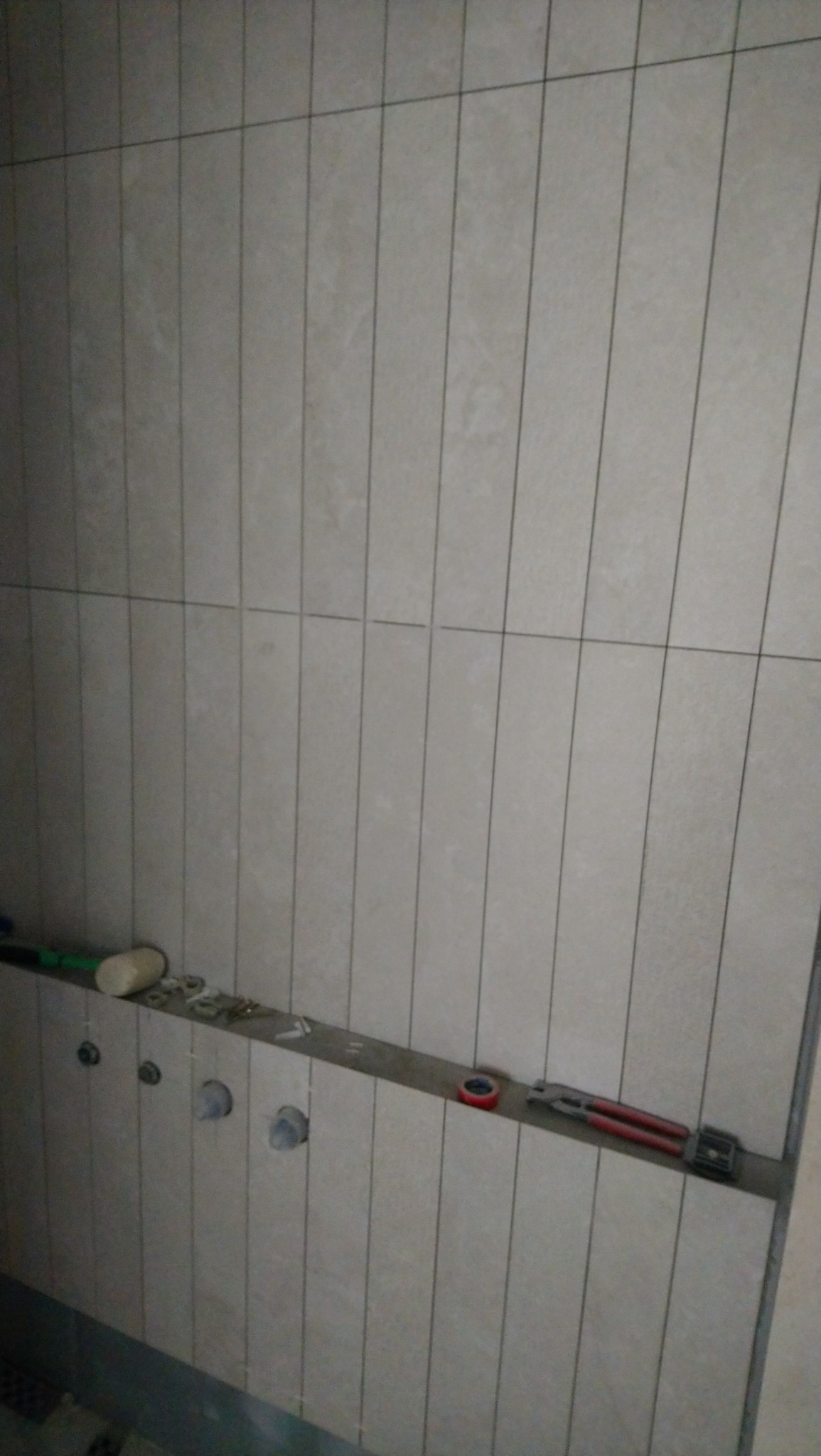 Ściana łazienki w trakcie wykańczania z ułożonymi pionowo jasnymi płytkami, widoczne narzędzia i elementy instalacji hydraulicznej.
