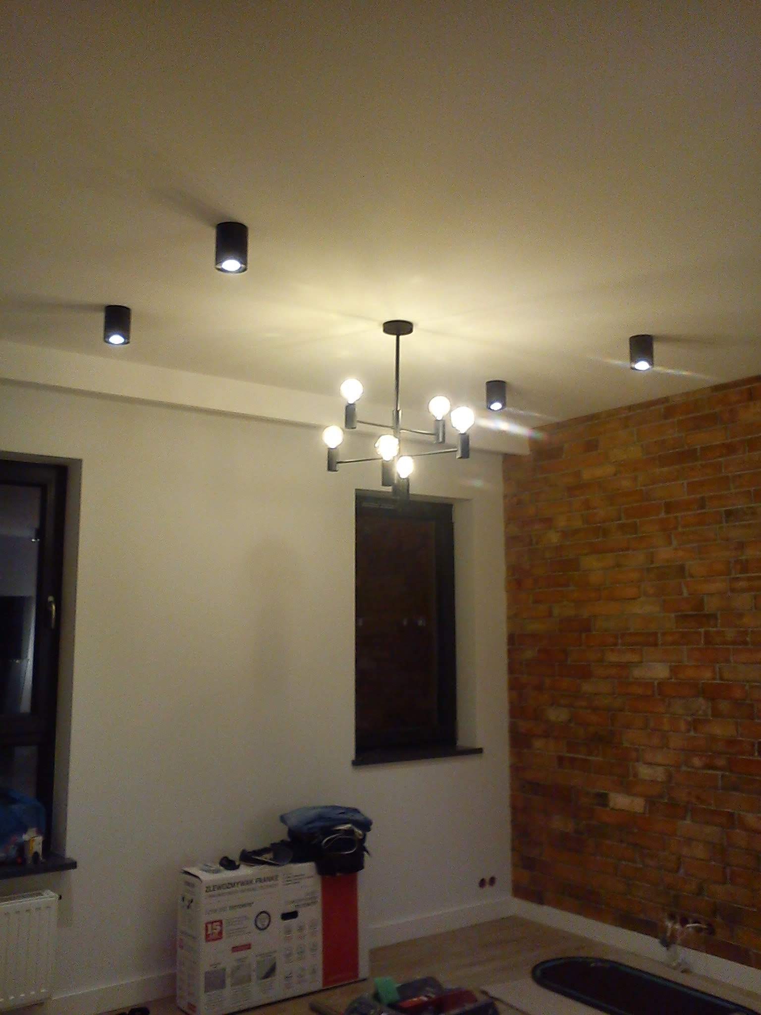 Wnętrze pomieszczenia po odświeżeniu w Wołominie, z nową lampą sufitową w stylu industrialnym i punktowymi reflektorami. Na ścianie widoczna dekoracyjna cegła, a obok okna stoi karton po zlewozmywaku.