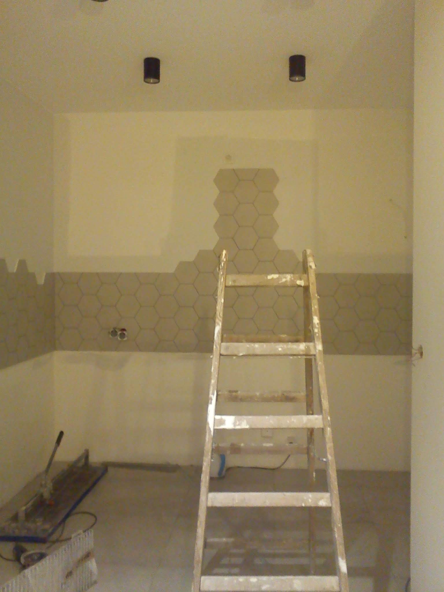 Łazienka w trakcie wykańczania z częściowo położonymi heksagonalnymi płytkami na ścianie, widoczna drabina malarska i narzędzia do cięcia płytek.