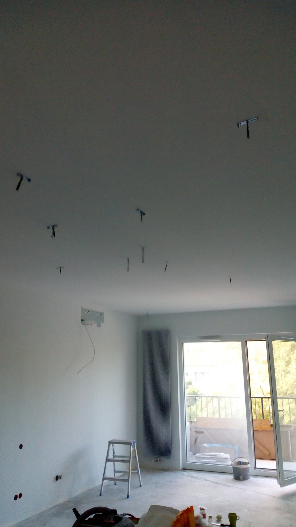 Wnętrze w trakcie remontu: widoczne kable wystające z sufitu, białe ściany, grzejnik, drabina, okno balkonowe z widokiem na zewnątrz.