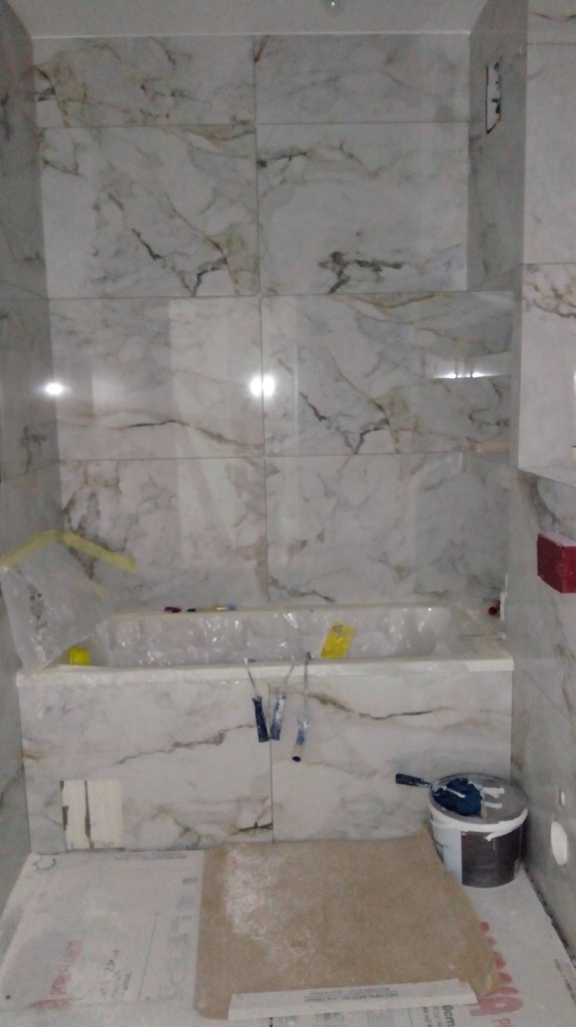 Wnętrze łazienki w trakcie prac wykończeniowych, widoczne marmurowe płytki na ścianach, zabezpieczona folią wanna, narzędzia malarskie i wiadro z farbą na podłodze.