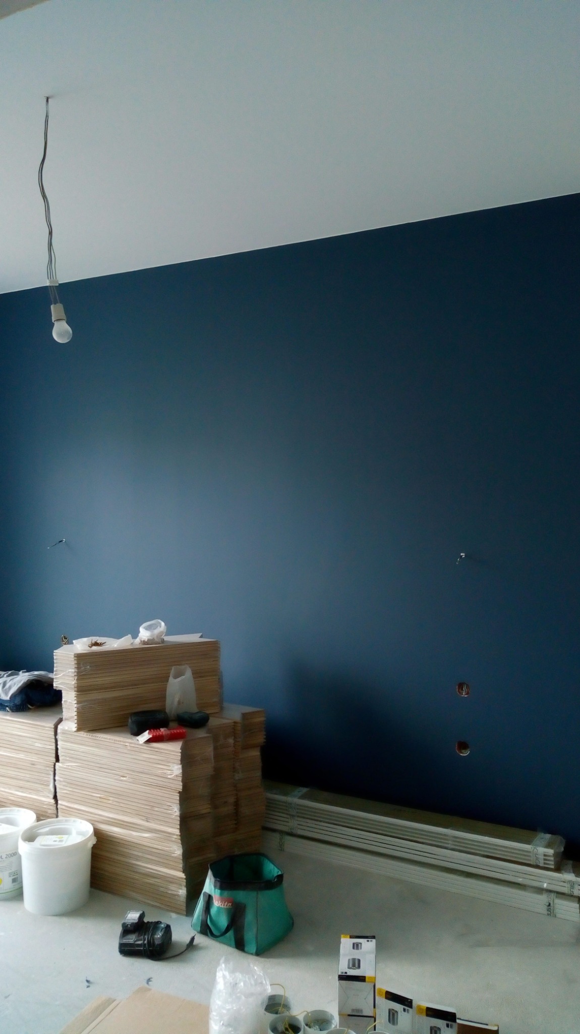 Świeżo pomalowana na granatowo ściana w pokoju, z widocznymi puszkami farby, stertą płyt karton-gips, torbą narzędziową Makita oraz listwami przypodłogowymi, pod wiszącą żarówką.