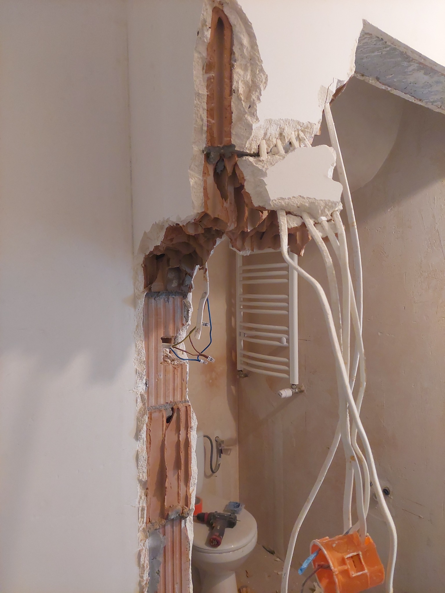 Demontaż ściany w łazience z odsłoniętymi przewodami elektrycznymi i rurami, widoczny grzejnik drabinkowy i toaleta, narzędzie na sedesie.