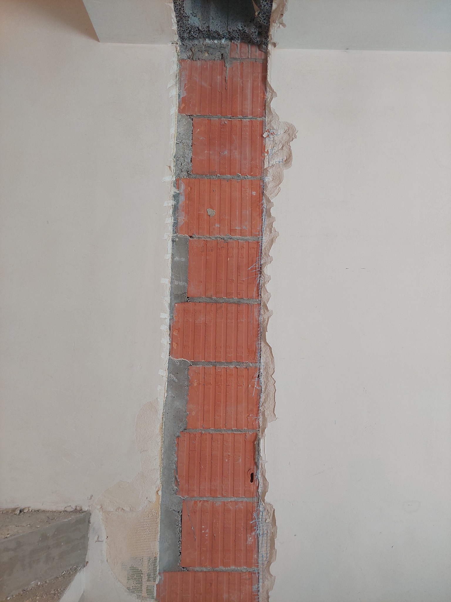 Wewnętrzna ściana z wyciętym pionowym fragmentem muru z czerwonej cegły, przygotowywana do wypełnienia i wyrównania, widoczne fragmenty siatki wzmacniającej.
