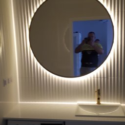 Kompleksowe Remonty Damilo dkk - Nowoczesna łazienka z okrągłym lustrem podświetlanym LED, umywalką z baterią w kolorze złotym i pralką w zabudowie.