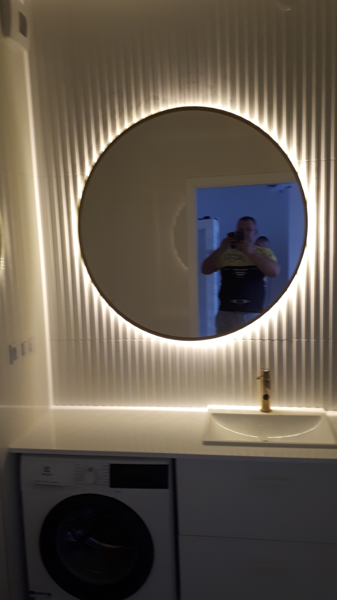 Nowoczesna łazienka z okrągłym lustrem podświetlanym LED, umywalką z baterią w kolorze złotym i pralką w zabudowie.