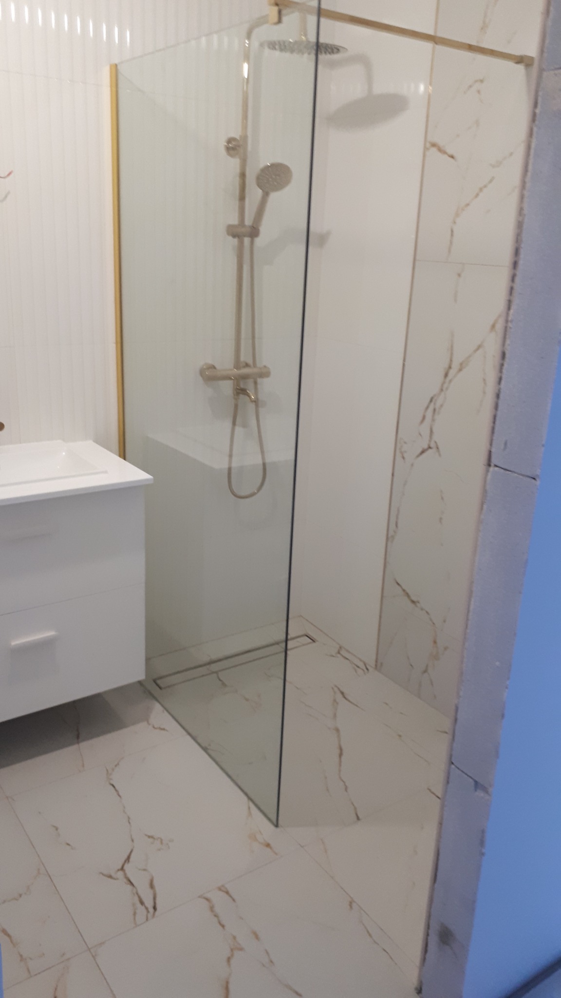 Nowoczesna kabina prysznicowa z złotymi elementami armatury i szklanymi ściankami, widoczna umywalka z szafką oraz marmurowe płytki podłogowe i ścienne.