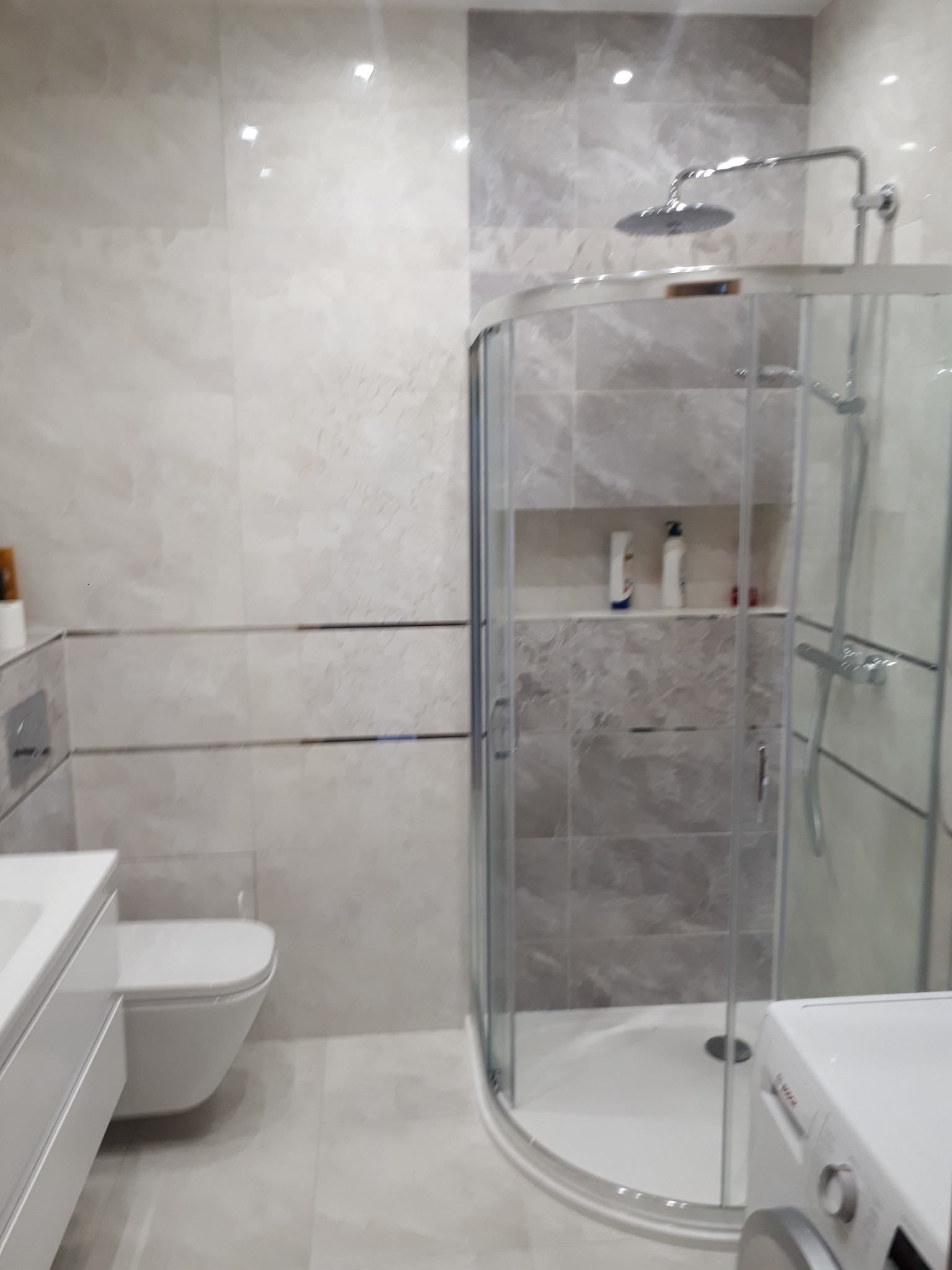 Nowoczesna łazienka z kabiną prysznicową typu walk-in, umywalką z szafką, podwieszaną toaletą i pralką, wykończona płytkami w odcieniach beżu i szarości z dekoracyjnymi listwami.