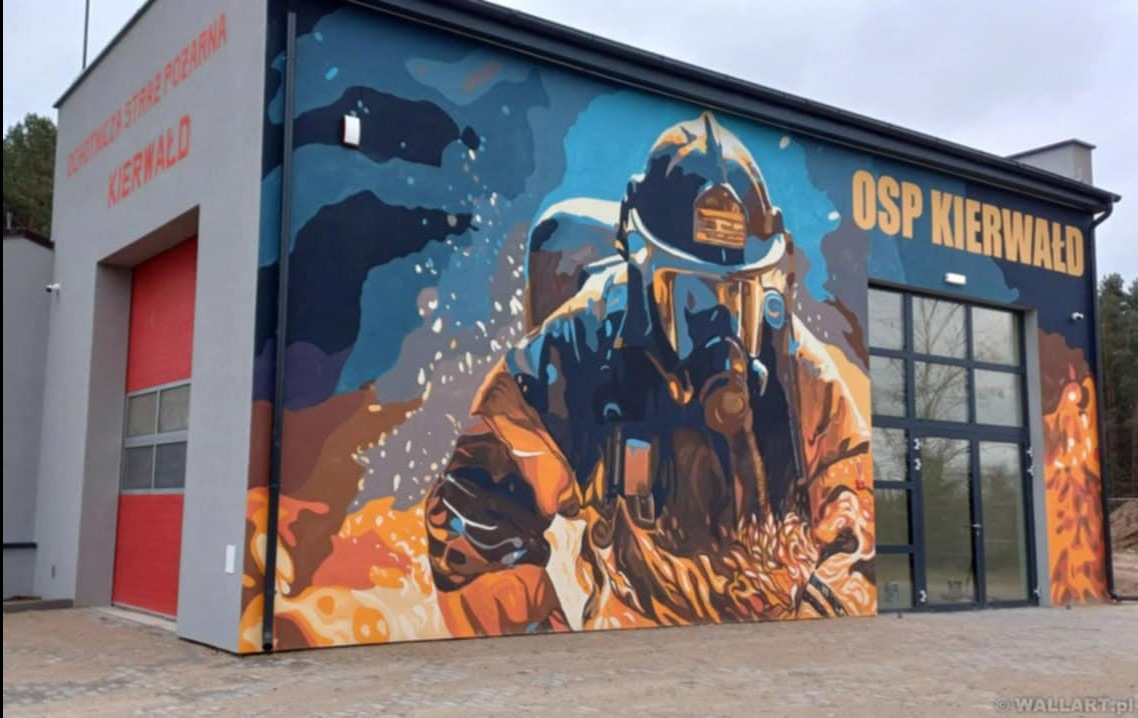Budynek OSP Kierwałd z muralem przedstawiającym strażaka w hełmie i masce, w dynamicznej kompozycji kolorów. Widoczne napisy i elementy architektoniczne budynku.