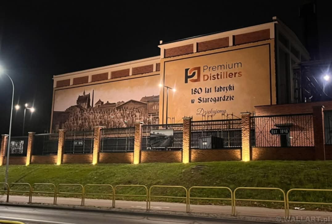 Mural z logo Premium Distillers na ścianie budynku, upamiętniający 180 lat fabryki w Starogardzie, oświetlony nocą, widoczny z ulicy.