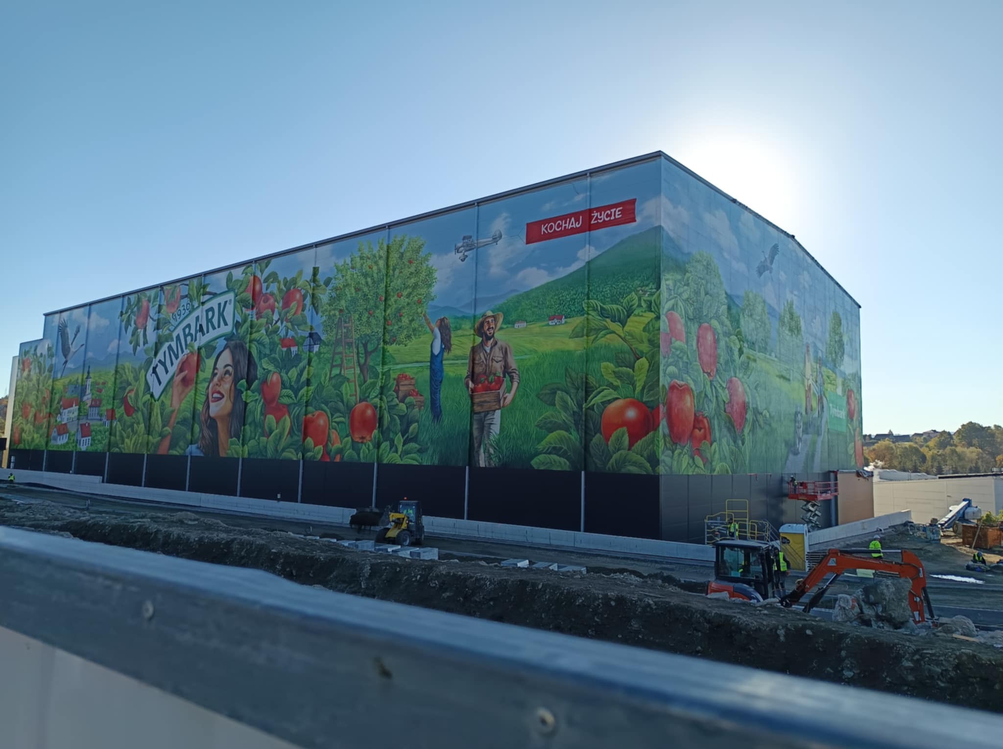 Ogromny mural reklamowy Tymbark na ścianie budynku, przedstawiający krajobraz z ludźmi i owocami, z widocznymi maszynami budowlanymi w tle.