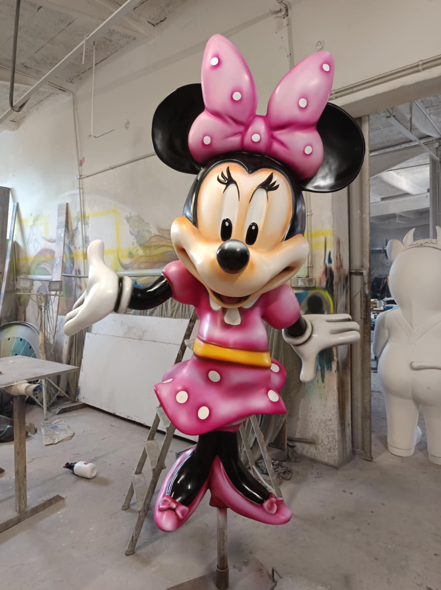 Figurka Myszki Minnie w różowej sukience i kokardą, wykonana z tworzywa sztucznego, w atelier artystycznym. Widoczne farby i drabina.