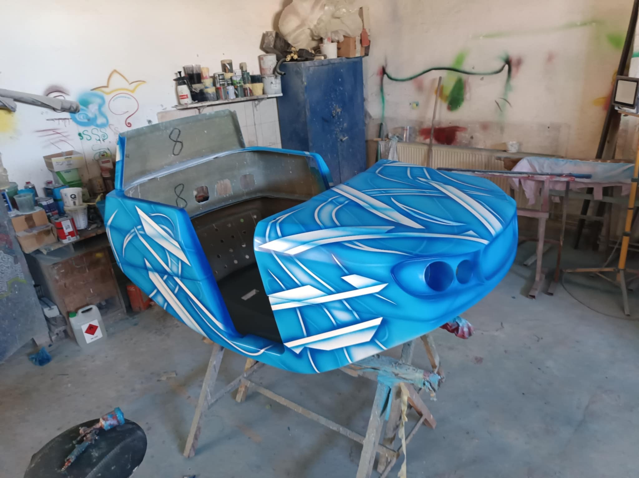 Błękitny kadłub pojazdu z białymi zdobieniami, umieszczony na drewnianym stojaku w warsztacie z graffiti na ścianach i narzędziami w tle. Detal malowania.