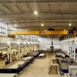 Wnętrze hali produkcyjnej z suwnicą i formami do odlew&oacute;w betonowych płyt, widoczni pracownicy w odblaskowych kamizelkach.