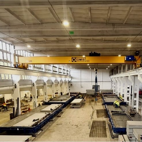 Wnętrze hali produkcyjnej z suwnicą i formami do odlewów betonowych płyt, widoczni pracownicy w odblaskowych kamizelkach.