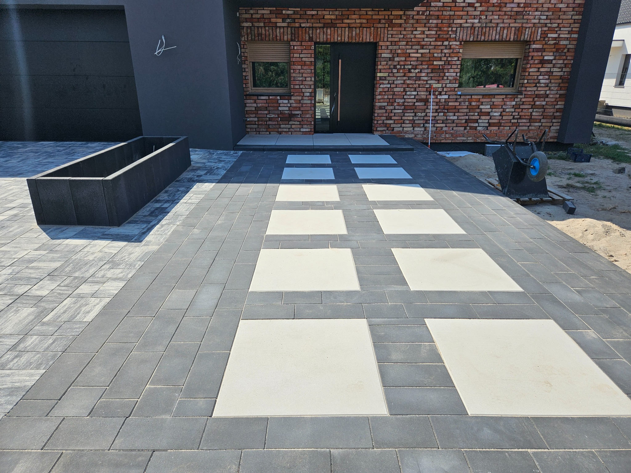 Nowoczesny podjazd z szarej kostki brukowej z wstawkami z jasnych płyt, prowadzący do domu z ceglaną elewacją i czarnymi elementami wykończenia, na drugim planie taczka.