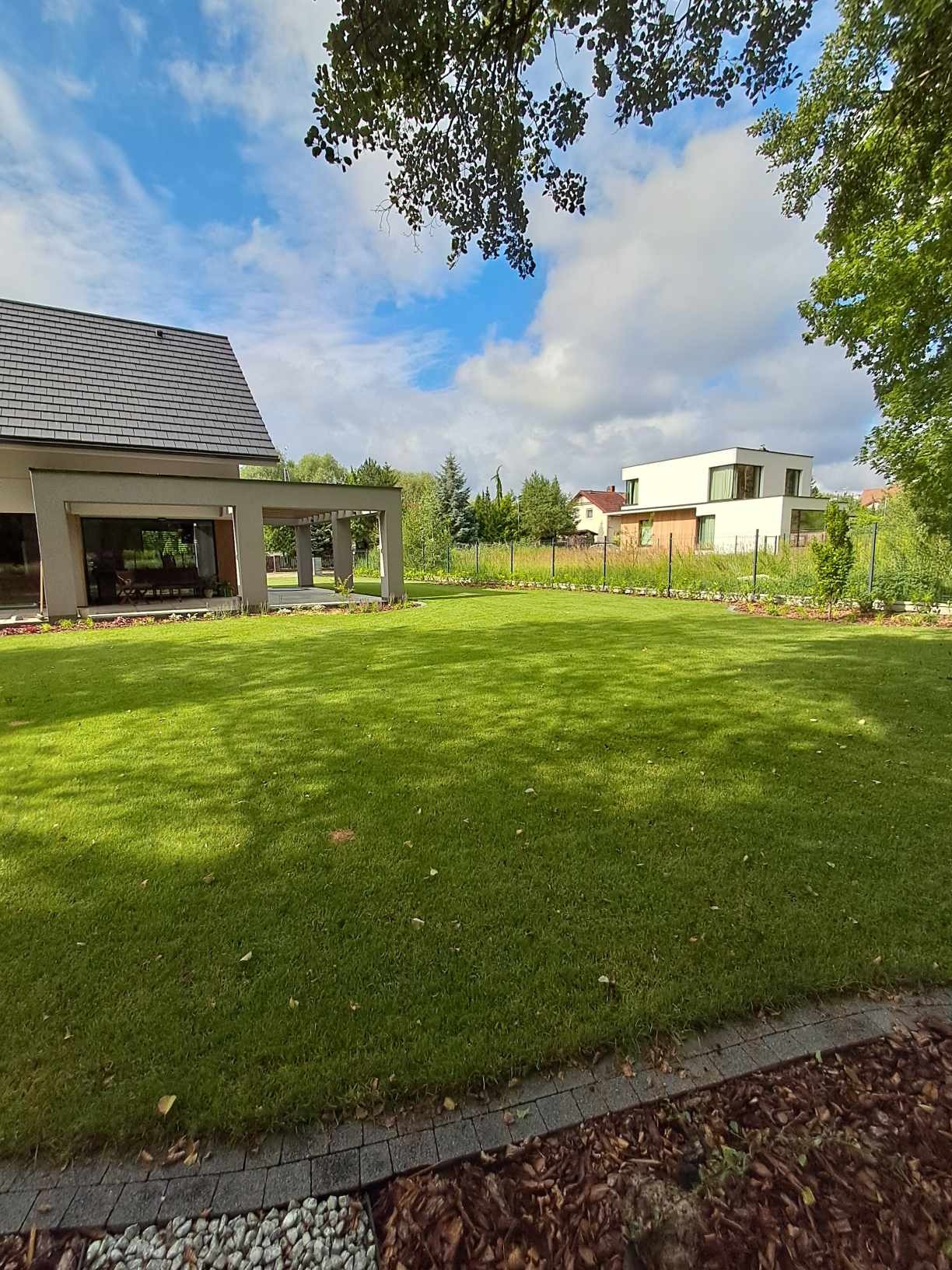 Rozległy, równo przystrzyżony trawnik przed nowoczesnym domem z szarą dachówką, widok z poziomu gruntu na tle błękitnego nieba z chmurami, obrzeże z kostki brukowej i kory.