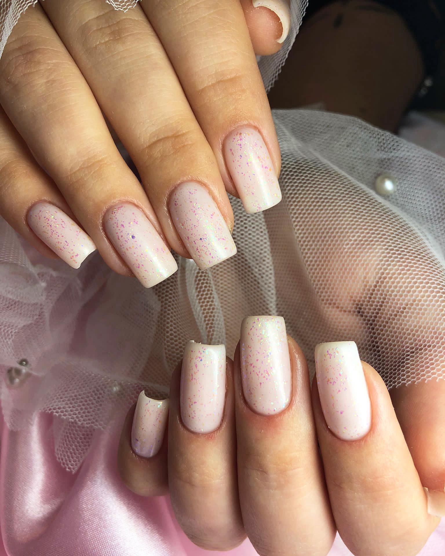 Delikatne, kwadratowe paznokcie w kolorze jasnego różu z opalizującymi drobinkami, prezentowane na tle białej koronki i różowego jedwabiu.