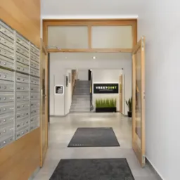 Wnętrze budynku biurowego z rzędem skrzynek pocztowych po lewej stronie, otwartymi drewnianymi drzwiami prowadzącymi do holu z logo firmy Vrestpoint i schodami w tle.