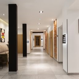 Vrestpoint - Nowoczesny korytarz biurowy z elementami cegły, czarnymi kolumnami, minimalistycznymi obrazami i szarą sofą z rośliną doniczkową.