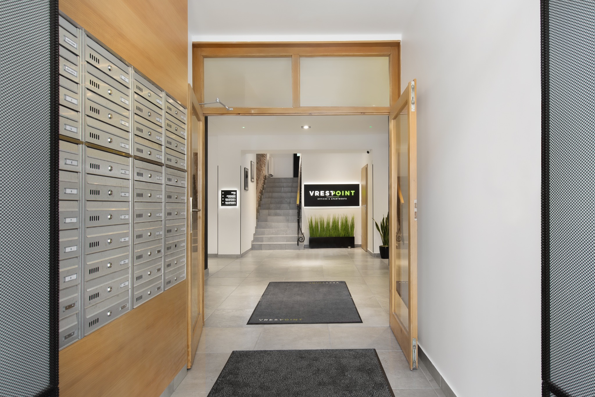 Wnętrze budynku biurowego z rzędem skrzynek pocztowych po lewej stronie, otwartymi drewnianymi drzwiami prowadzącymi do holu z logo firmy Vrestpoint i schodami w tle.