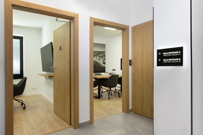 Wnętrze biura z trzema otwartymi drzwiami do sal konferencyjnych oznaczonych literami A i B, widoczny stół konferencyjny, krzesła biurowe i czarno-biała fotografia miasta na ścianie.