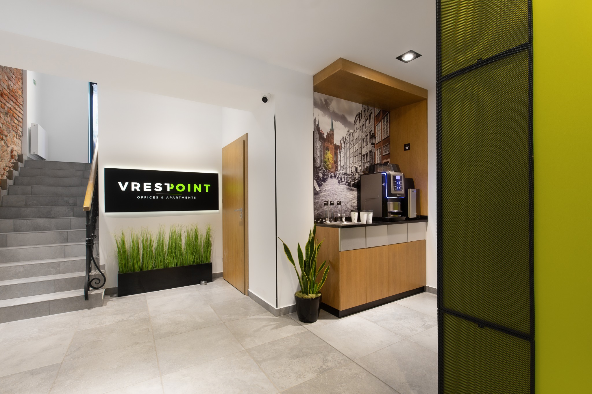 Nowoczesne wnętrze biurowe z recepcją, schodami z ceglaną ścianą i automatem do kawy na tle fototapety z motywem miejskim.