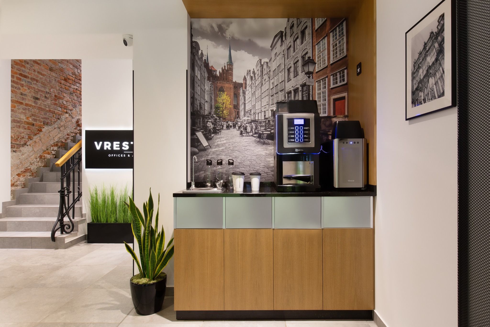 Nowoczesna przestrzeń biurowa w Gdańsku z aneksem kuchennym: ekspres do kawy na blacie z drewnianymi szafkami, w tle fototapeta z motywem gdańskiej uliczki i schody z roślinami doniczkowymi.