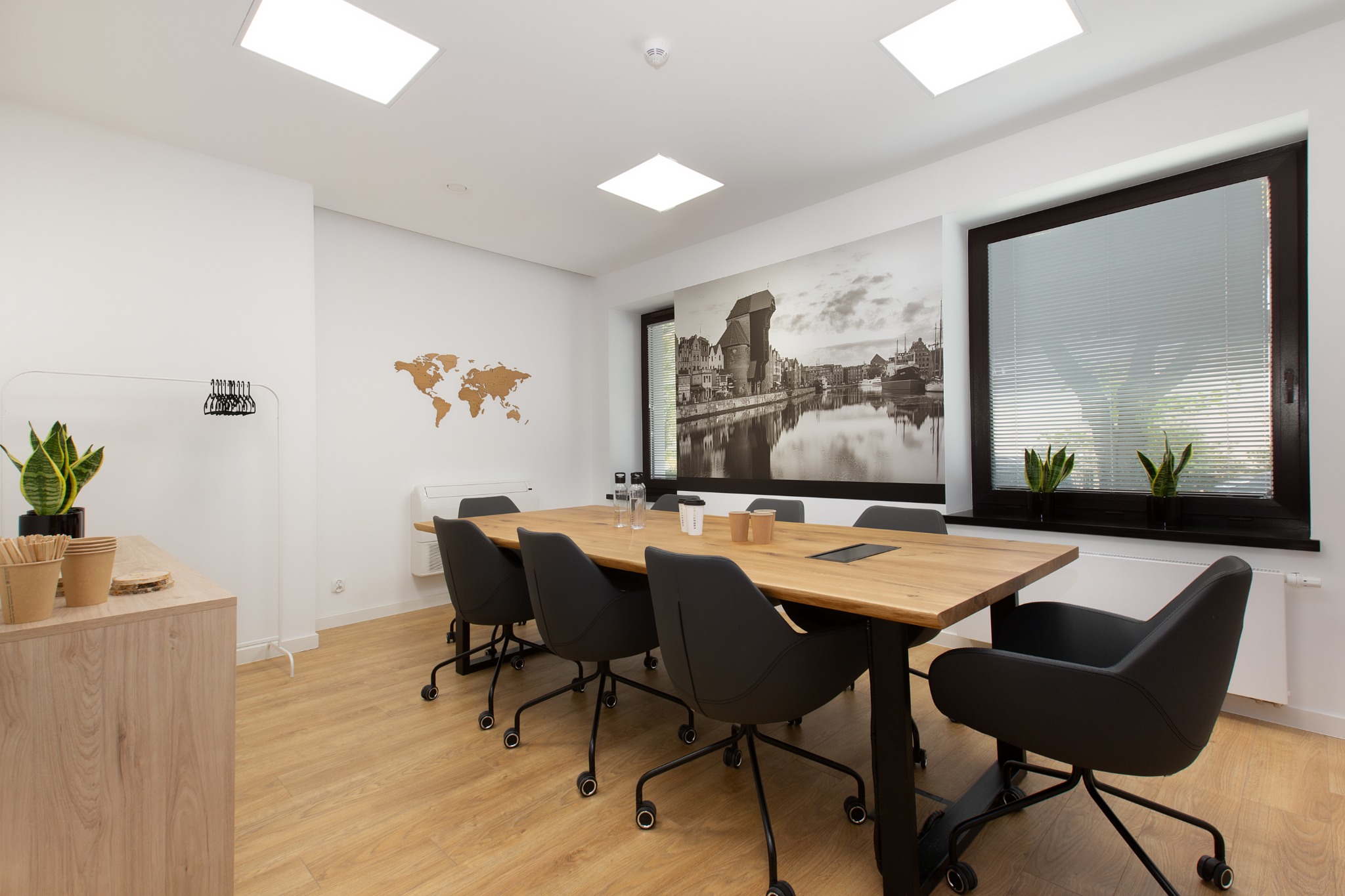 Jasne, minimalistyczne wnętrze biurowe z drewnianym stołem konferencyjnym, czarnymi krzesłami na kółkach, mapą świata na ścianie i fototapetą z motywem miasta nad rzeką.