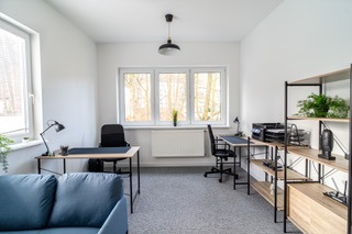 Jasne, przestronne wnętrze biurowe z dwoma biurkami, krzesłami, regałem z roślinami i szarą wykładziną, oświetlone naturalnym światłem z trzech okien.